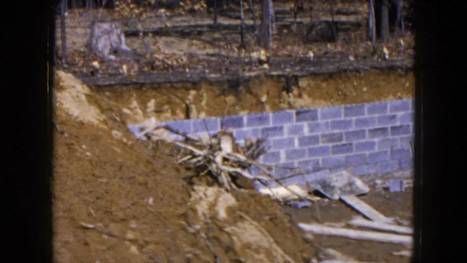 1954 Hicksville frame showing a partially laid gray block foundation wall set into a dirt excavation with boards and debris nearby.