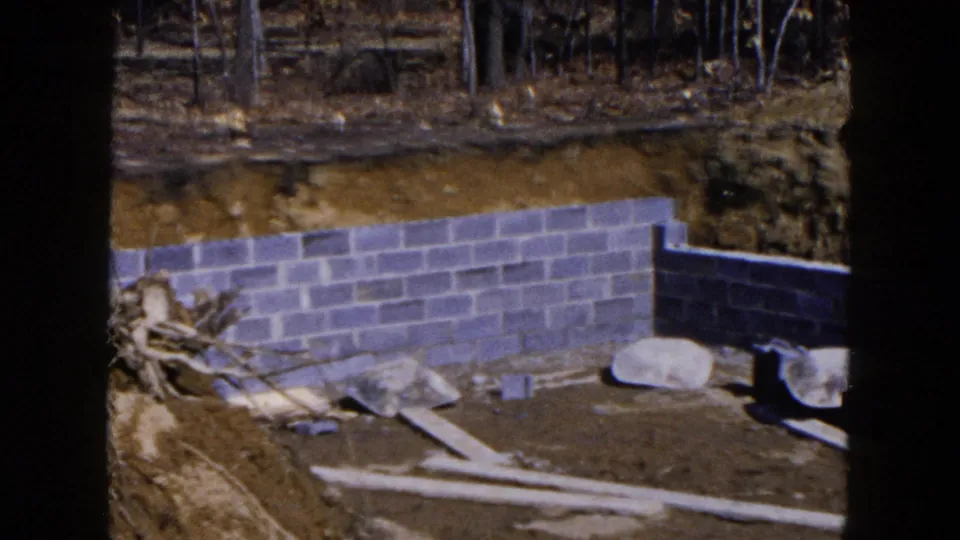 1954 Hicksville frame with a cinder-block basement corner, excavated soil, loose planks, stones, and trees at the edge of the lot.