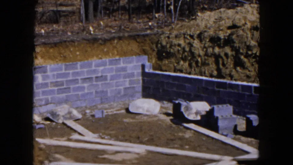 1954 Hicksville construction frame showing a partially built cinder-block foundation, loose blocks, large stones, planks, and excavated earth.