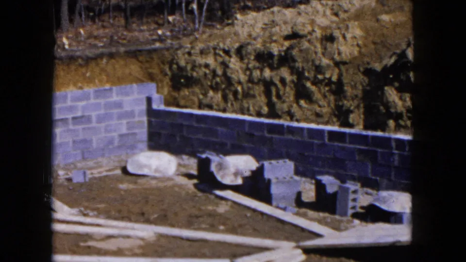 1954 Hicksville frame showing the foundation corner, stacked loose blocks, stones, boards, excavated soil, and woods beyond the site.