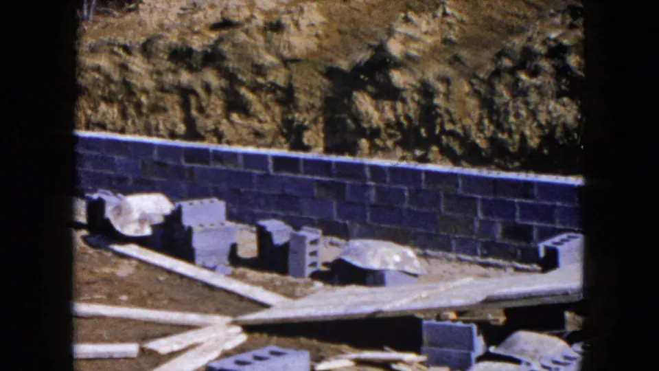 A 1954 Hicksville homebuilding view with cinder blocks, a foundation wall, loose boards, and earthwork behind it.