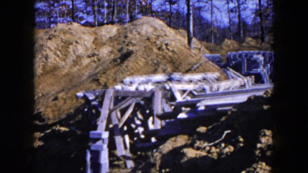 Construction site with dirt piles, wooden formwork, and stacked concrete blocks near bare trees.