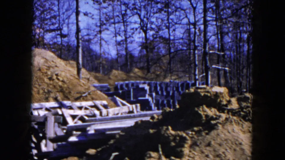 Outdoor residential construction detail in Hicksville, showing wood framing, stacked blocks, earth piles, and a wooded edge.