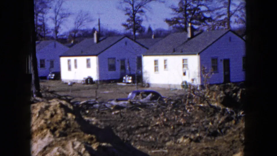 Completed white houses in Hicksville, New York, seen across muddy construction ground with parked cars and bare trees.