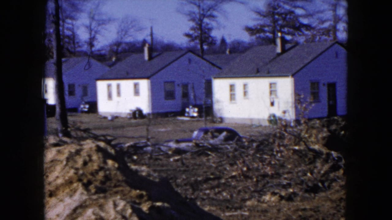 1954 Hicksville home-movie frame showing small white homes behind graded earth, bare trees, and a partly visible car.
