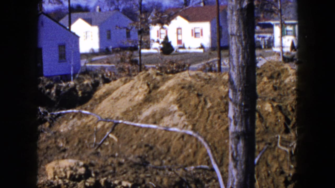 Residential trench with soil piles, bare trees, utility poles, and small houses beyond.