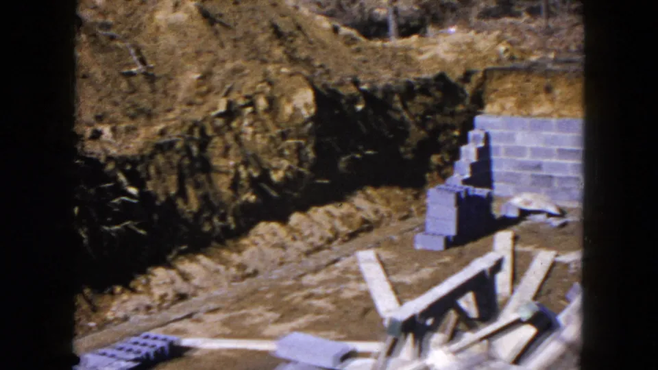 A partially built cinder-block foundation wall stands in a trench with stacked blocks and wooden beams nearby.