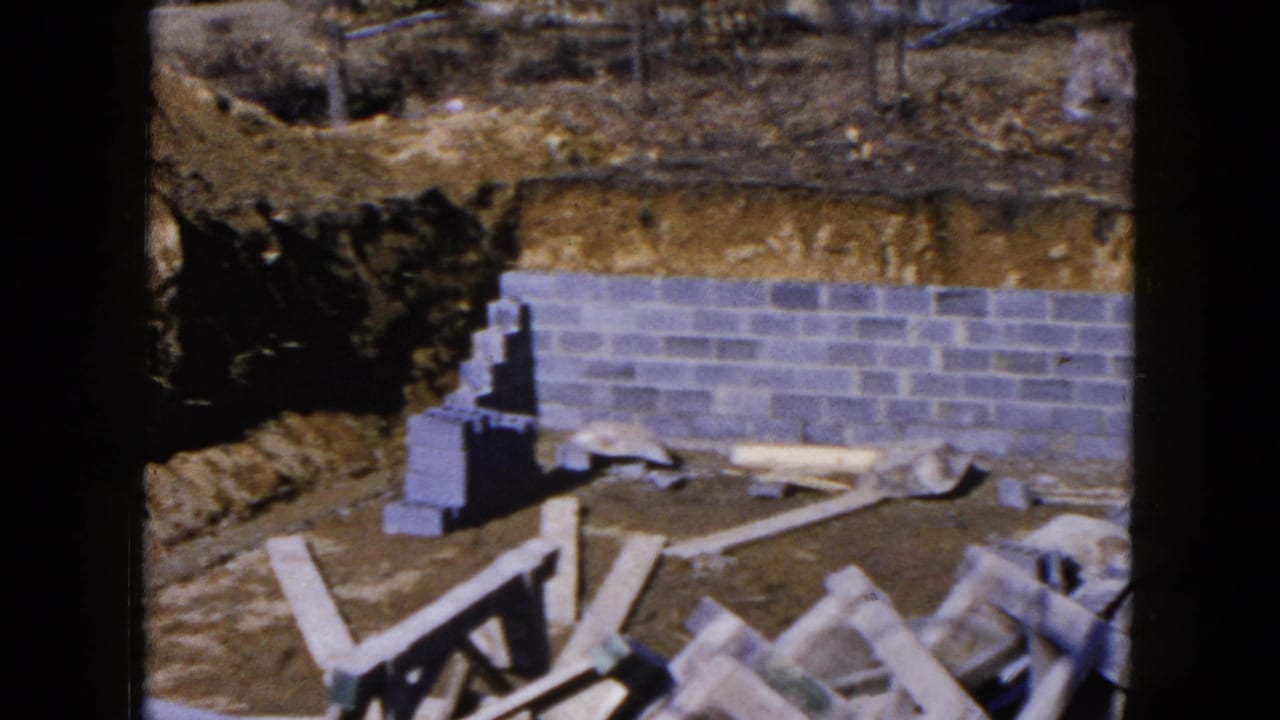 1954 Hicksville home-movie frame showing a gray cinder block foundation wall set into a brown dirt excavation with scattered construction materials.
