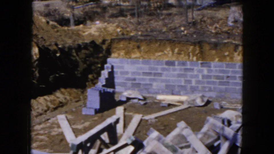 1954 Hicksville home-movie frame showing a gray cinder block foundation wall set into a brown dirt excavation with scattered construction materials.