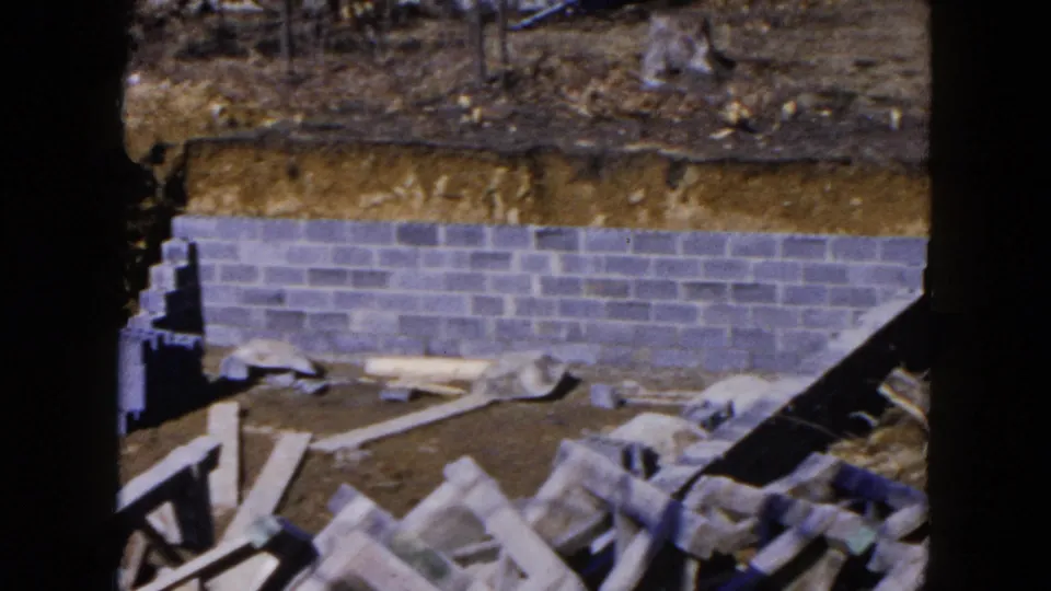 1954 Hicksville construction view with a cinder block foundation, exposed soil, scattered boards, and masonry materials seen through a dark frame.