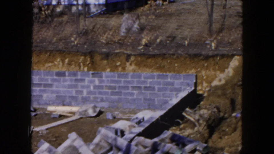 Home-movie frame from Hicksville in 1954 showing a cinder block foundation below a rough dirt slope with boards and bare trees around the lot.