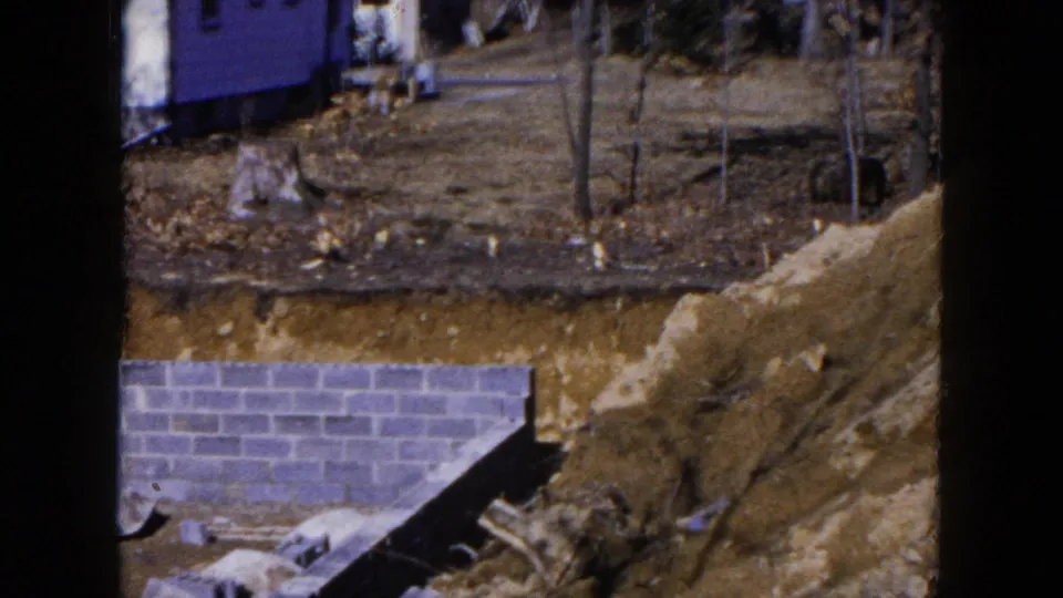 1954 Hicksville frame showing a partially built cinder block foundation, large dirt piles, bare trees, and a small background structure.