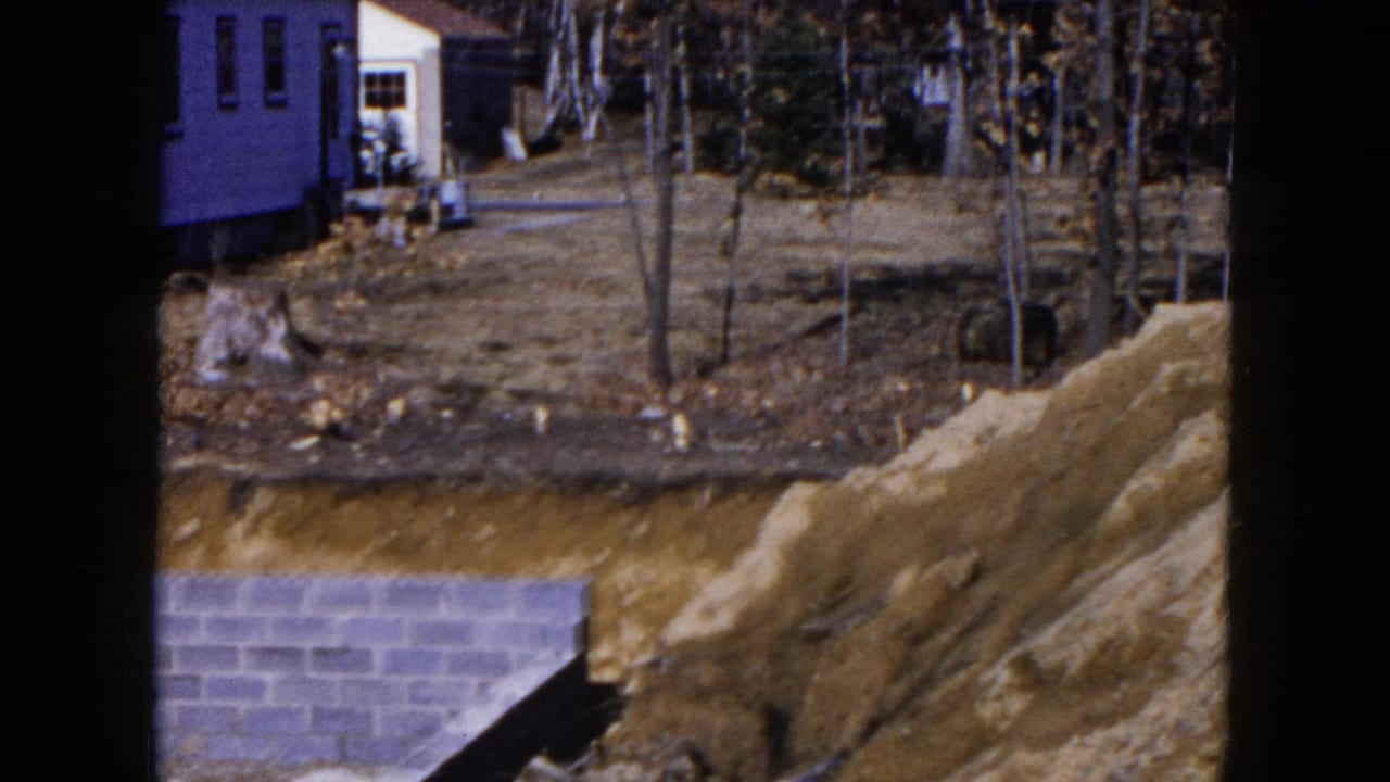 1954 Hicksville 8mm frame showing a partly built house site with dirt banks, trees, and nearby residential structures.