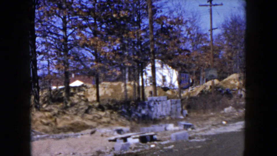 1950s 8mm view of a Hicksville roadside construction area with woodland, concrete blocks, dirt, and houses beyond.