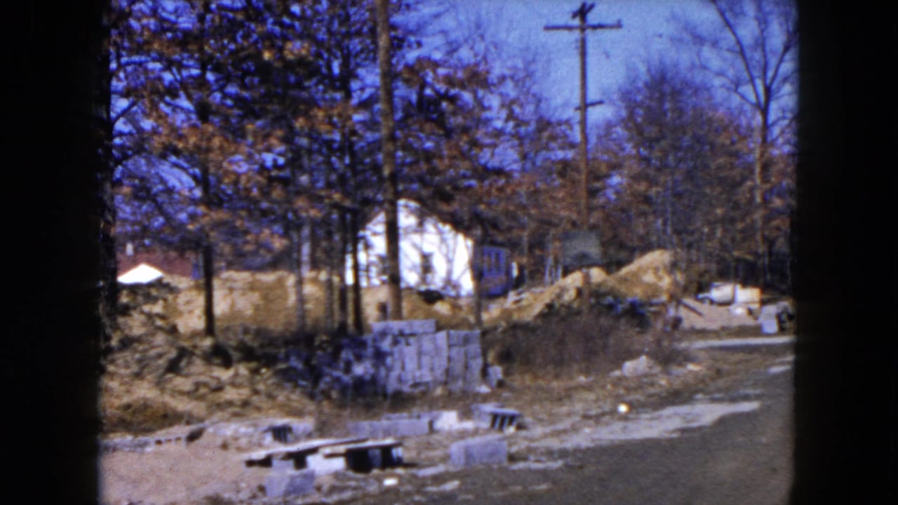 Roadside construction materials beside trees, utility poles, and a white house in the background.