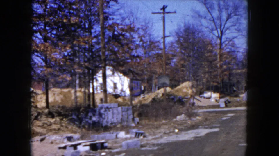 1954 Hicksville construction site seen from the road, with sand piles, concrete blocks, utility poles, and a white house partly screened by trees.