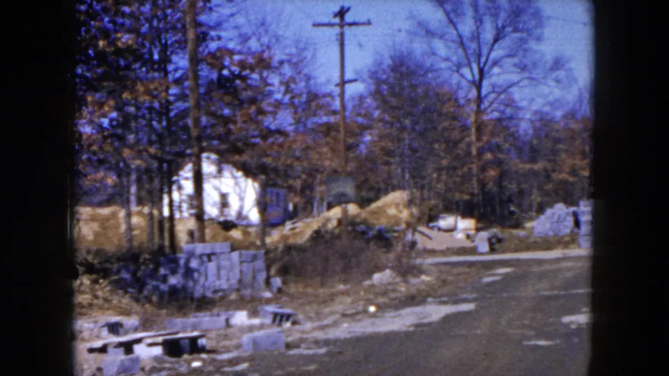 Hicksville, New York construction area in 1954 with a curving road, block stacks, sand piles, utility poles, and a white house behind trees.
