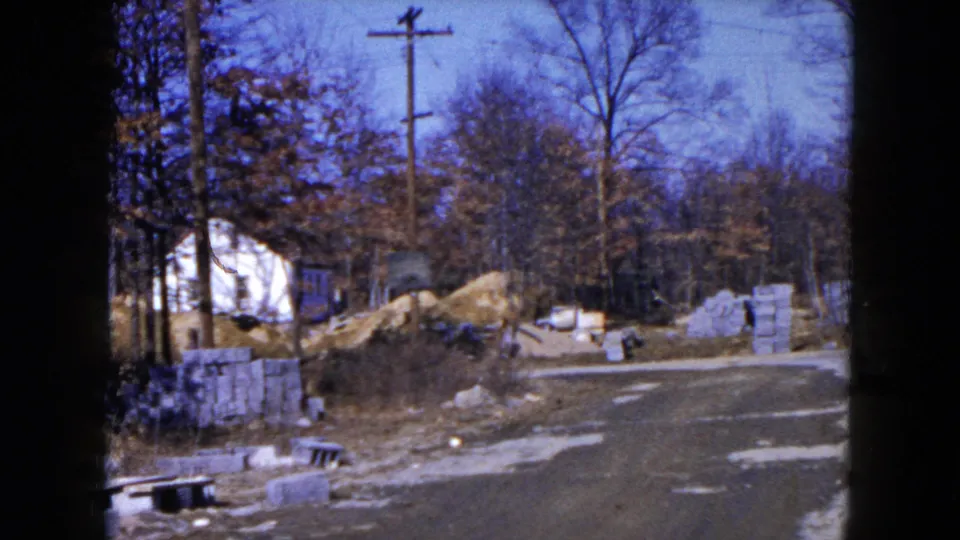 Final 1954 Hicksville frame showing roadside construction materials, a large sand pile, bare trees, utility poles, and a white house in the background.