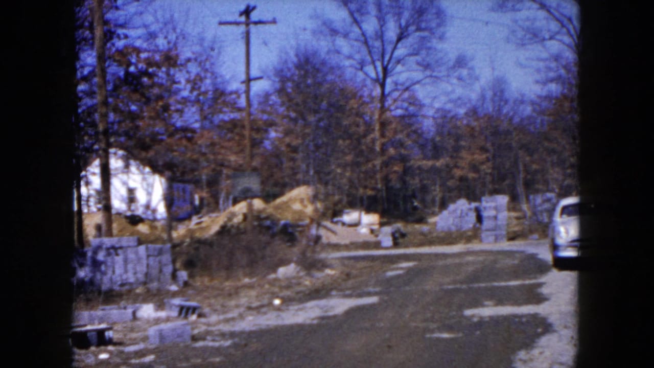 1954 Hicksville home-movie frame showing a gravel road, stacked blocks, dirt piles, bare trees, and part of a light blue car.