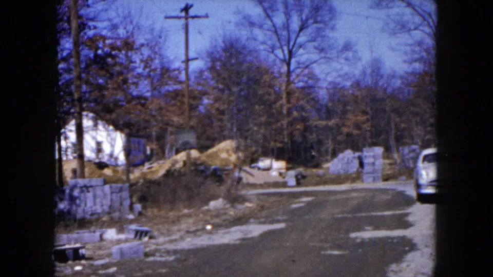 1954 Hicksville home-movie frame showing a gravel road, stacked blocks, dirt piles, bare trees, and part of a light blue car.