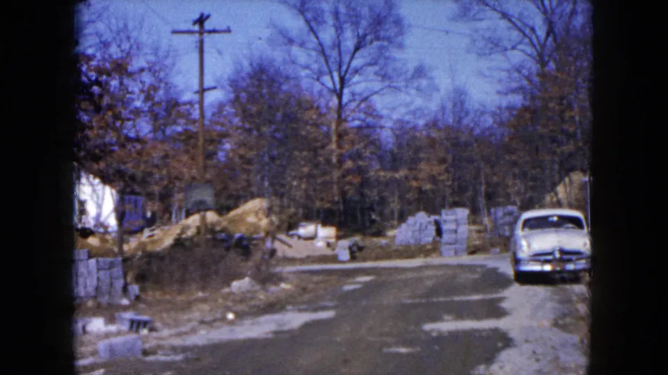 1954 Hicksville frame with a light blue car parked near a gravel road, concrete blocks, dirt piles, and leafless trees.