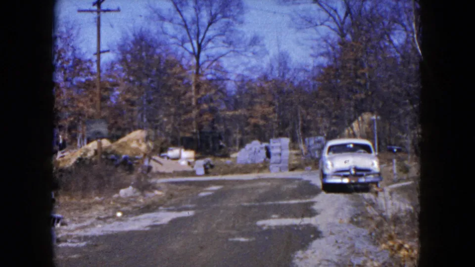 1954 Hicksville home-movie frame showing a parked light blue car beside a road with construction blocks and bare trees.