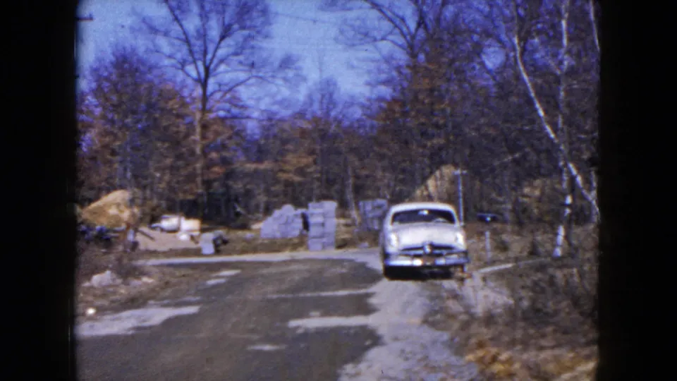 1954 Hicksville frame of a light blue car parked along a quiet road near concrete blocks, brush, and leafless trees.
