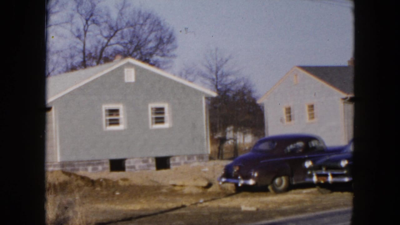 Gray single-family house beside a dark 1950s sedan on an unpaved residential street.
