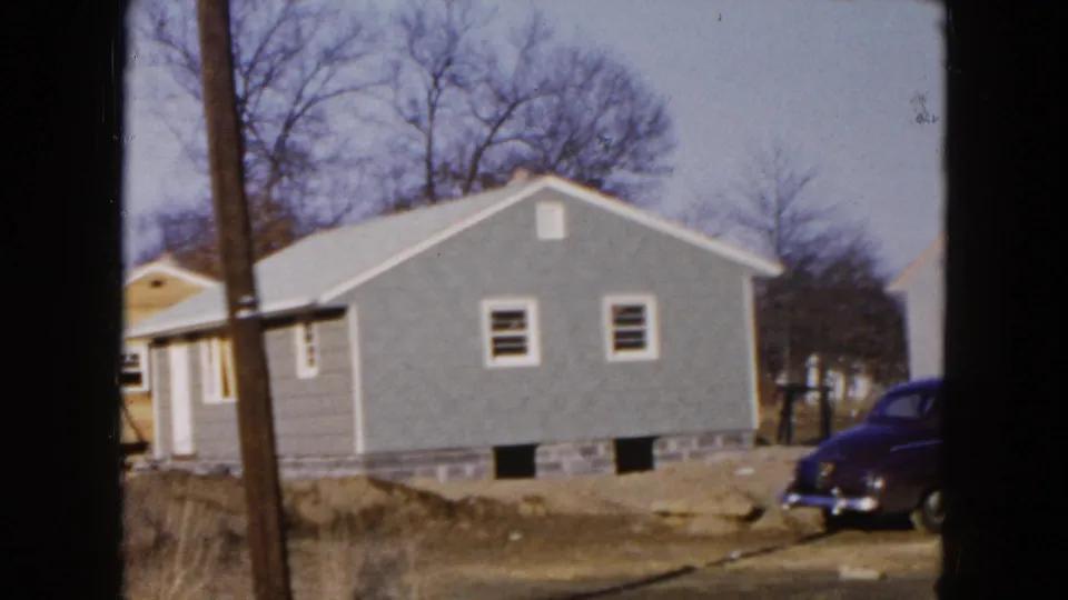 1954 Hicksville residential view with a gray house, utility pole, bare trees, and the rear of a dark 1950s car at right.