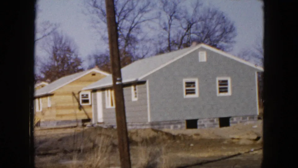 1954 Hicksville home-movie frame showing adjacent gray and light wood-sided houses, a utility pole, and leafless trees.