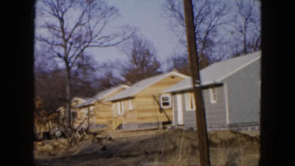 1954 Hicksville home-movie frame showing yellow and blue suburban houses with bare trees, muddy ground, and a foreground utility pole.