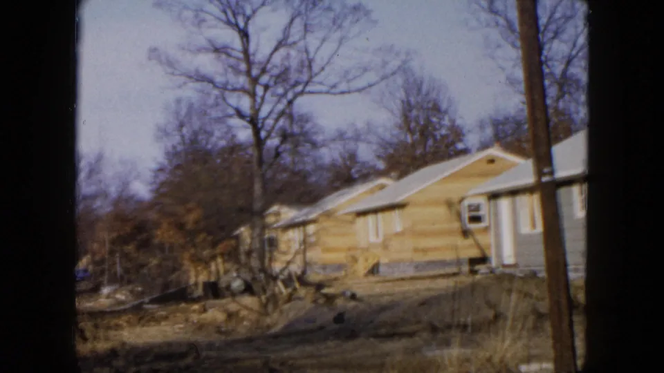 1954 Hicksville frame with a yellow house and blue side wall beyond rough winter ground and leafless trees.