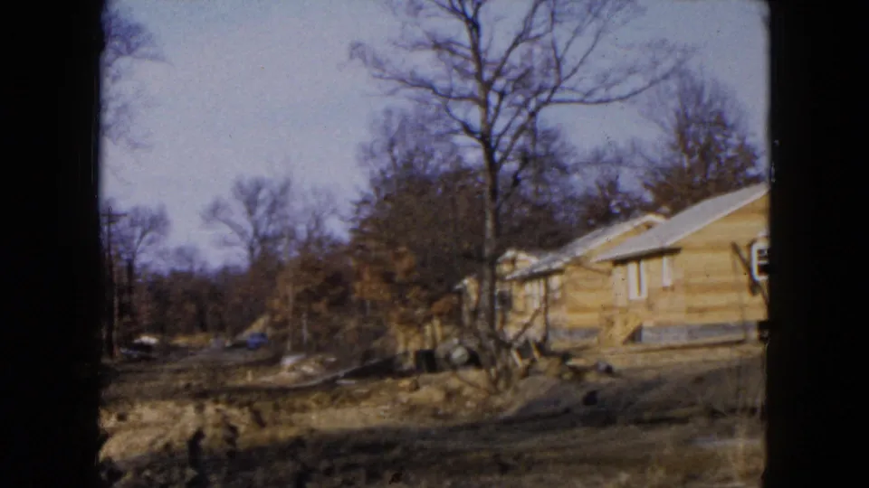 1954 Hicksville home-movie frame showing a yellow house at the edge of the shot with bare trees and undeveloped ground.