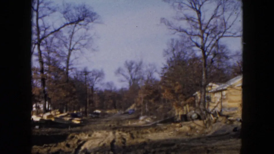 1954 Hicksville frame showing open undeveloped ground, leafless trees, and the partial edge of a yellow suburban house.