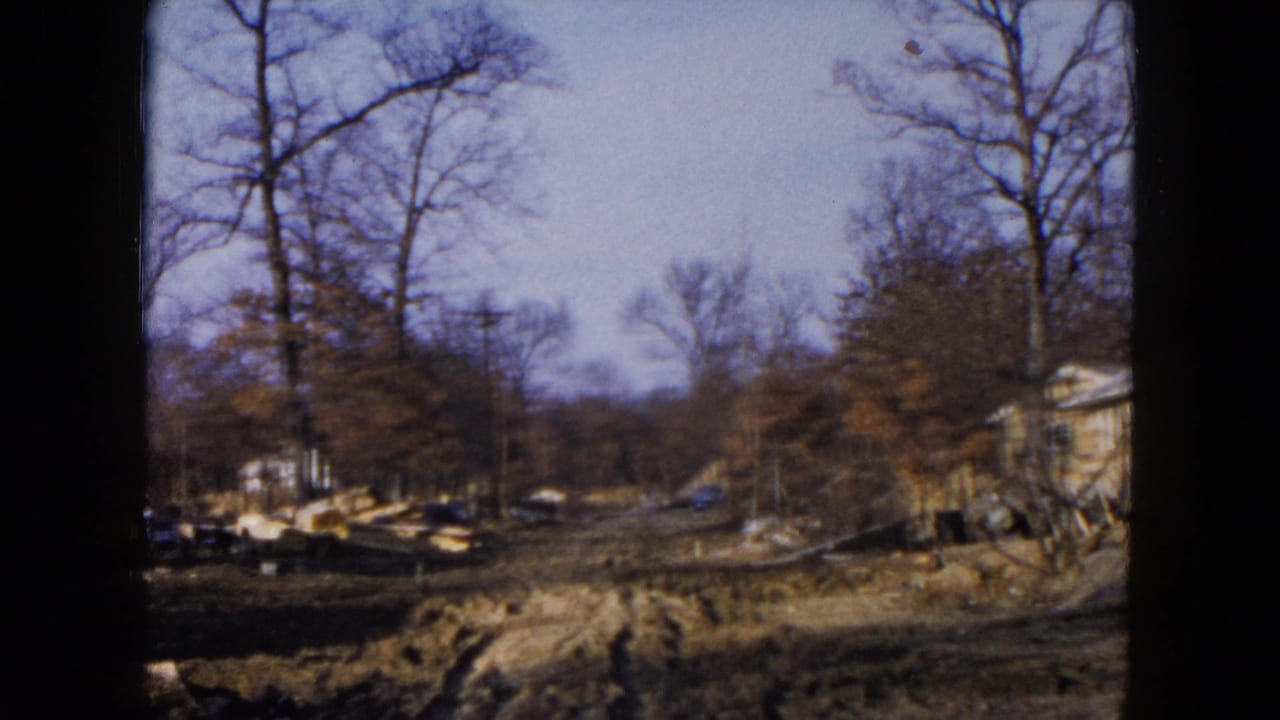 1954 Hicksville 8mm frame showing a cleared wooded house site with mud, lumber piles, bare trees, and a small structure at right.