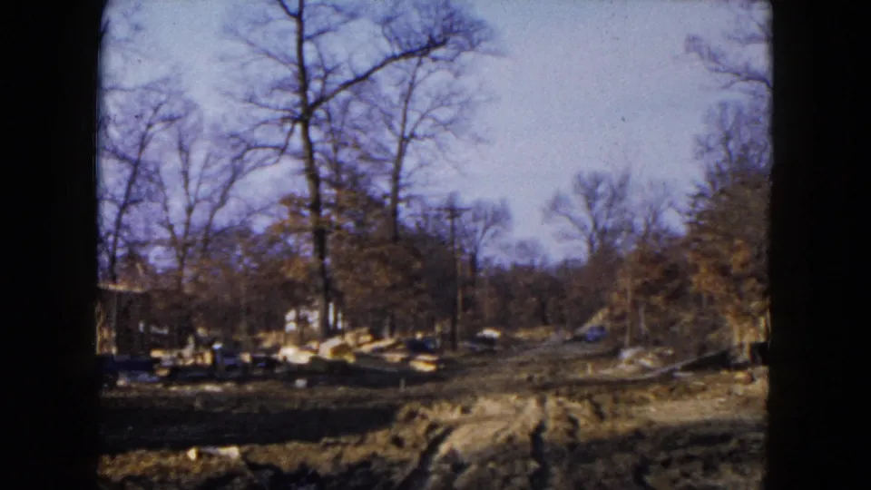 1954 Hicksville home-movie frame of a wooded suburban construction site with muddy ground, bare trees, lumber, and a house edge.