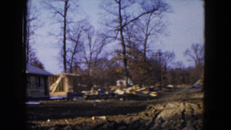 1954 Hicksville 8mm frame showing a partly built house in a wooded lot, with exposed framing and construction debris nearby.