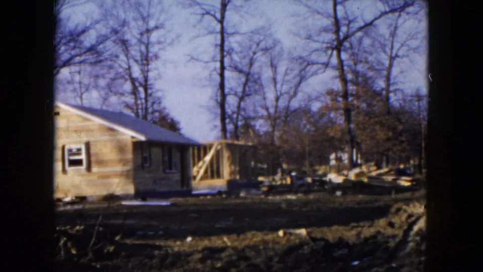 1954 Hicksville frame showing a small house under construction, with exposed porch framing, lumber, bare trees, and muddy ground.