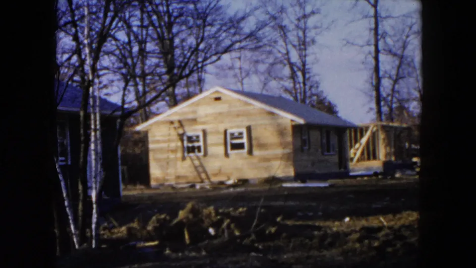 1954 Hicksville exterior view of a house being built, with a ladder against the wall and trees behind it.