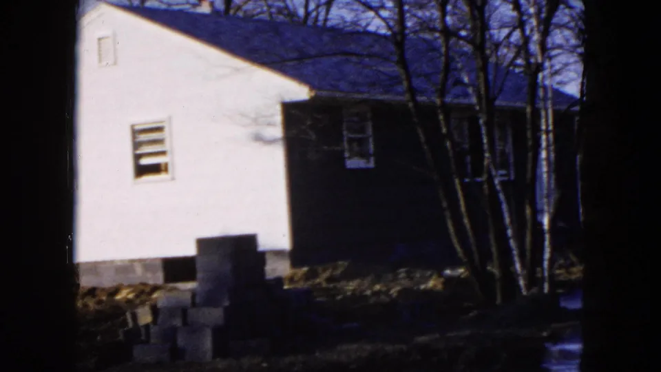 1954 Hicksville frame showing a nearby white house, stacked concrete blocks, trees, and a blue car at the lot edge.