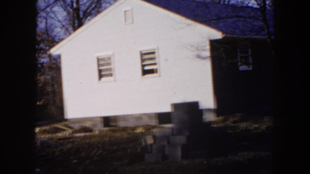 White single-story house beside a darker structure on a dirt lot with trees behind it.