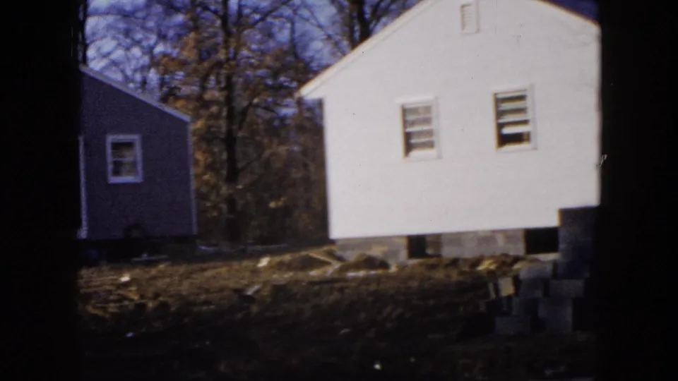 1954 Hicksville frame with a white house, gray side building, dark tree trunks, and scattered material on open ground.