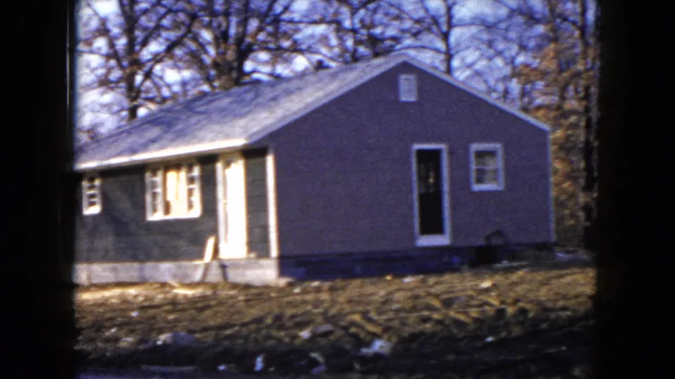 1954 Hicksville frame showing a gray single-story house with a dark doorway, windows, pale trim, bare trees, and rough ground.