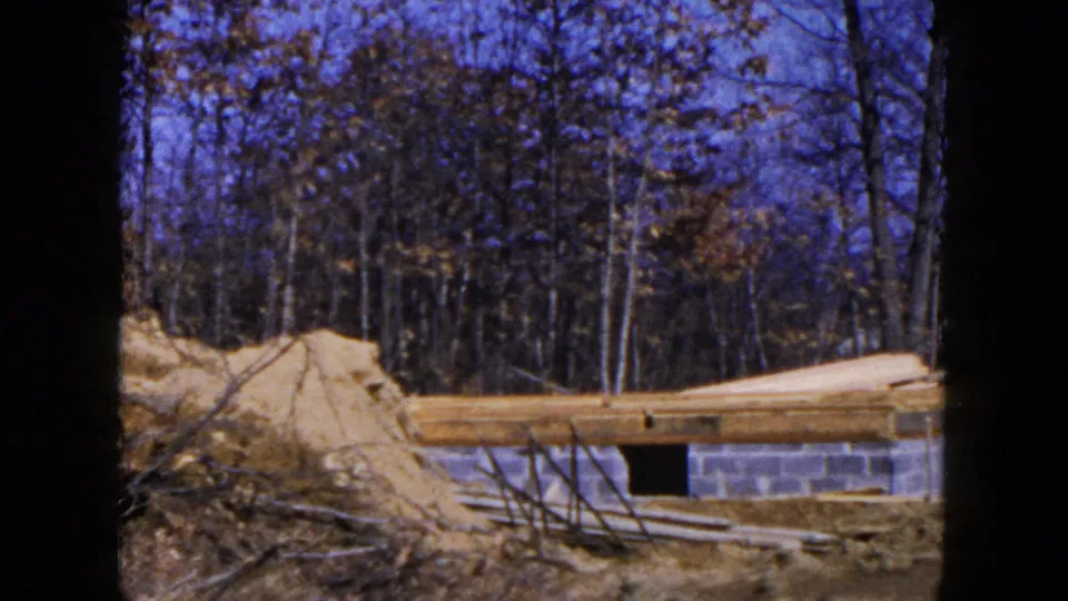 1954 Hicksville frame showing a concrete block foundation, long lumber beams, dirt piles, and leafless trees behind the site.