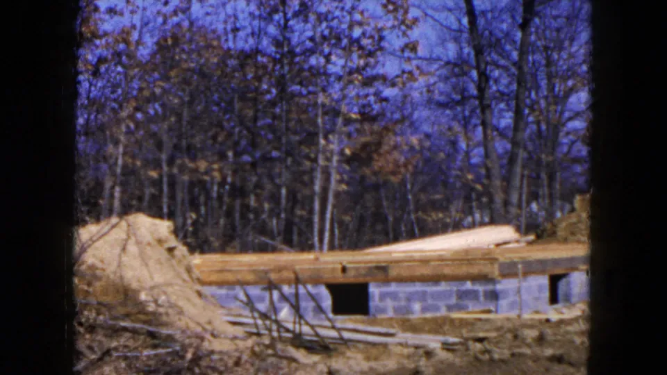 Home-movie view of lumber laid over a block foundation in a wooded Hicksville construction area, with dirt and branches in front.