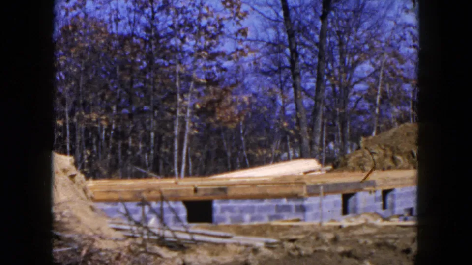 1954 Hicksville construction frame with long wood planks resting above concrete blocks, dirt banks, and leafless trees in the background.