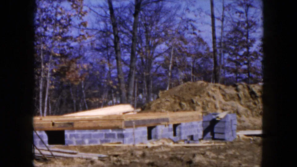 Mid-century 8mm frame of the Hicksville house construction site, with block walls, floor framing, lumber, and a large dirt pile.