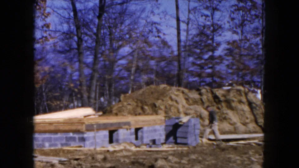 1954 Hicksville construction frame showing a worker near the right edge beside the foundation, lumber, dirt mound, and bare trees.