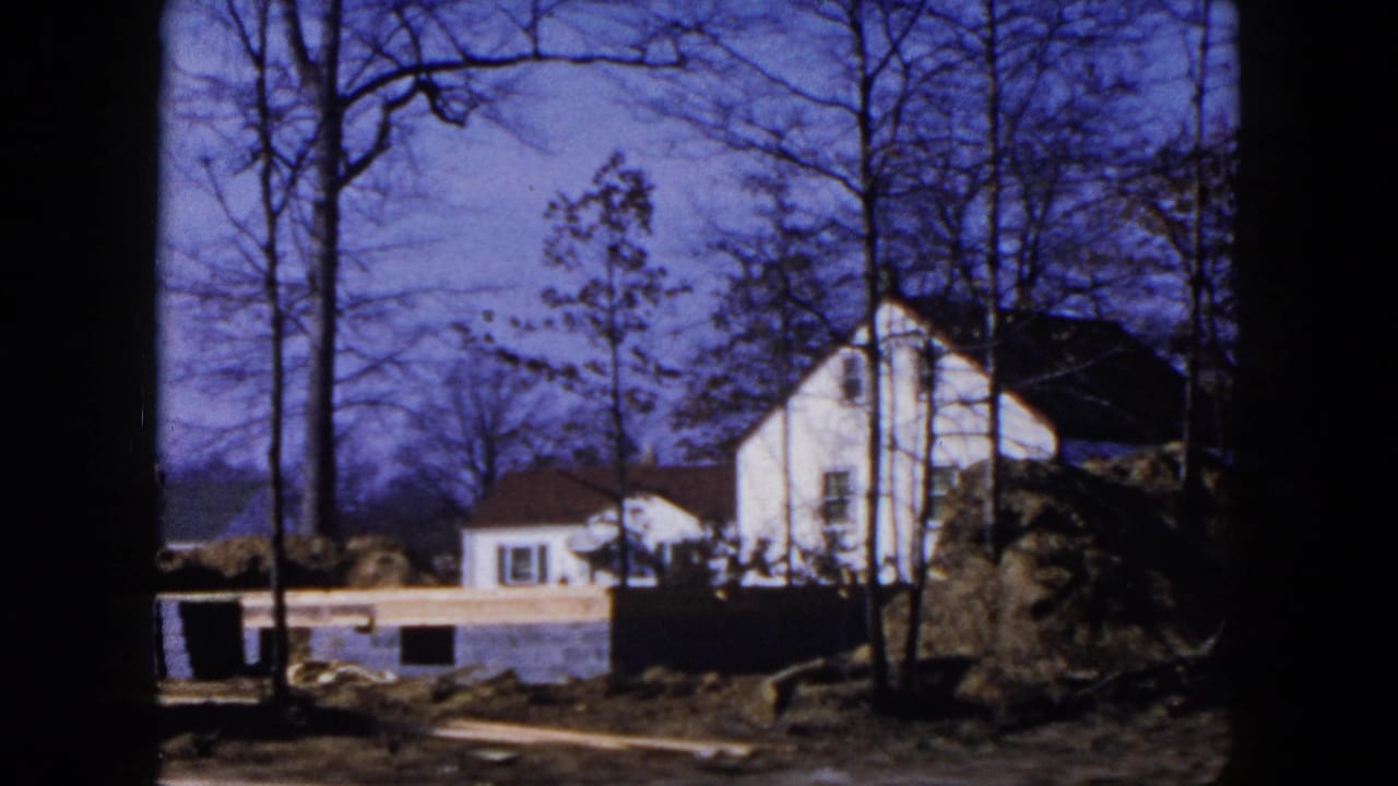 1954 Hicksville home-movie frame showing a white house, bare trees, a foundation wall, and earth piles at a suburban construction site.