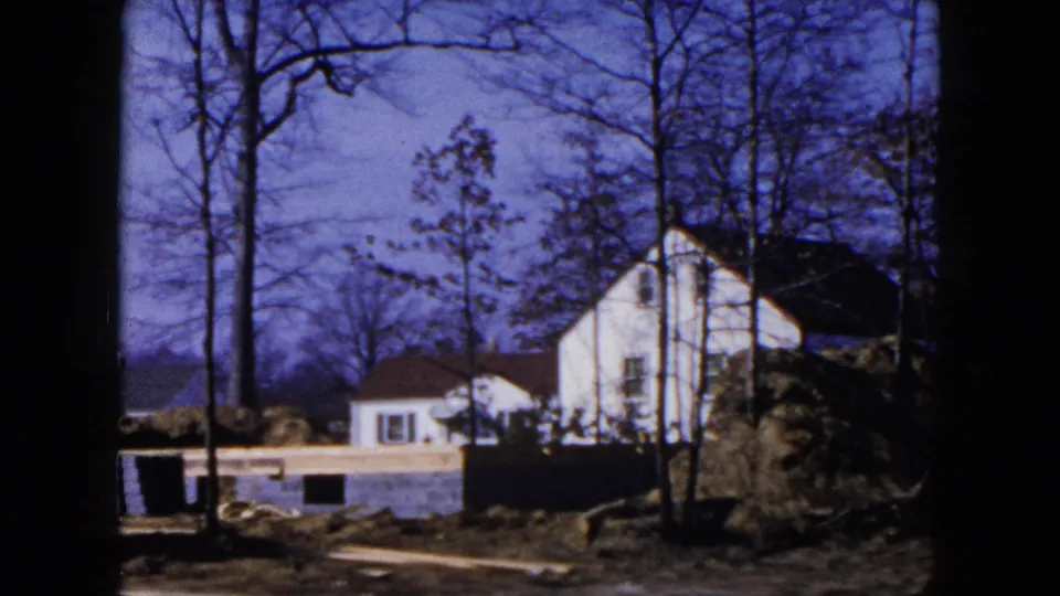 1954 Hicksville home-movie frame showing a white house, bare trees, a foundation wall, and earth piles at a suburban construction site.